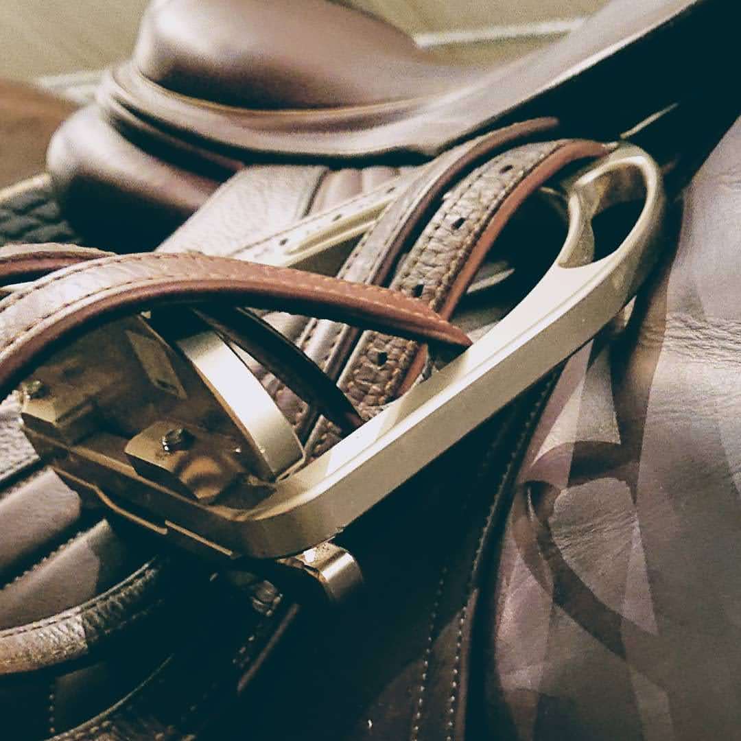 Eine Nahaufnahme eines braunen Ledersattels. Im Fokus steht ein moderner, silberner Sicherheitssteigbügel aus Metall, durch den die braunen Steigbügelriemen aus Leder gefädelt sind. Die Textur des gepflegten Leders und die technischen Details des Bügels sind deutlich erkennbar. Das Bild hat einen warmen, leicht matten Filter.