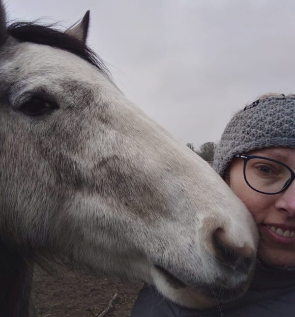 Nahaufnahme einer lächelnden Frau mit Brille und grauer Mütze, die ihren Kopf sanft an die Nüster eines weißen Schimmels lehnt